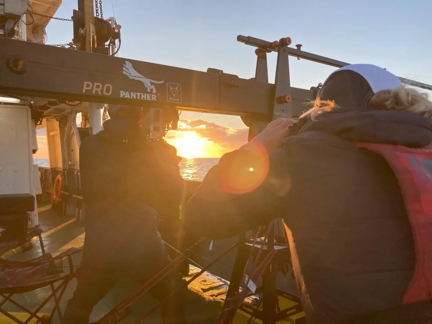 Camera crew filming from the deck of a ship at sunset, with a crane arm overhead and one person in a life jacket.