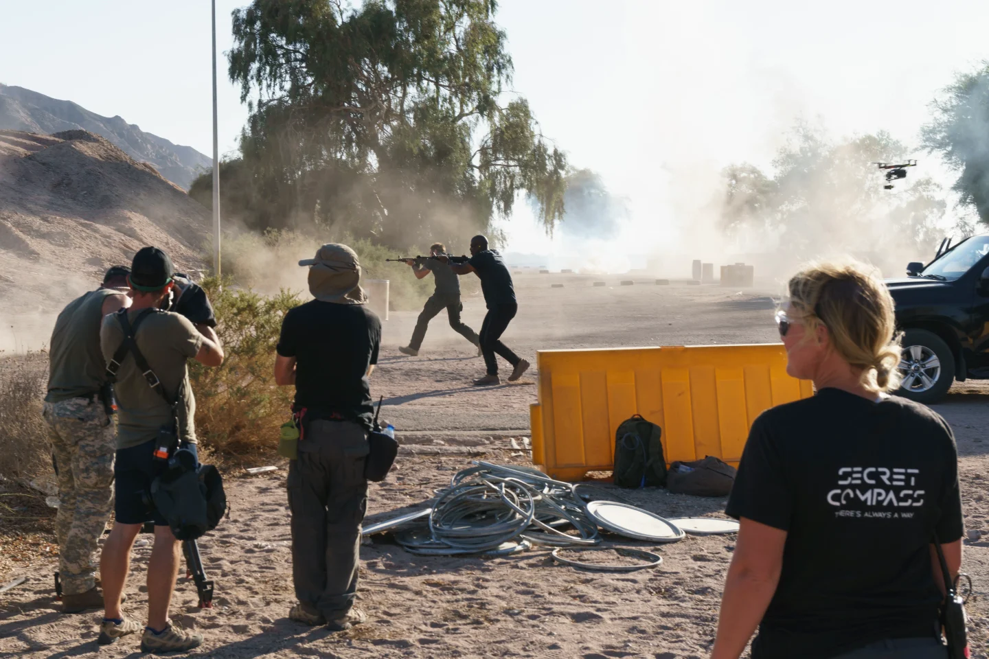 Secret Compass safety adviser watching from behind a barrier as crew film two actors with rifles in a dusty, smoke-filled scene.
