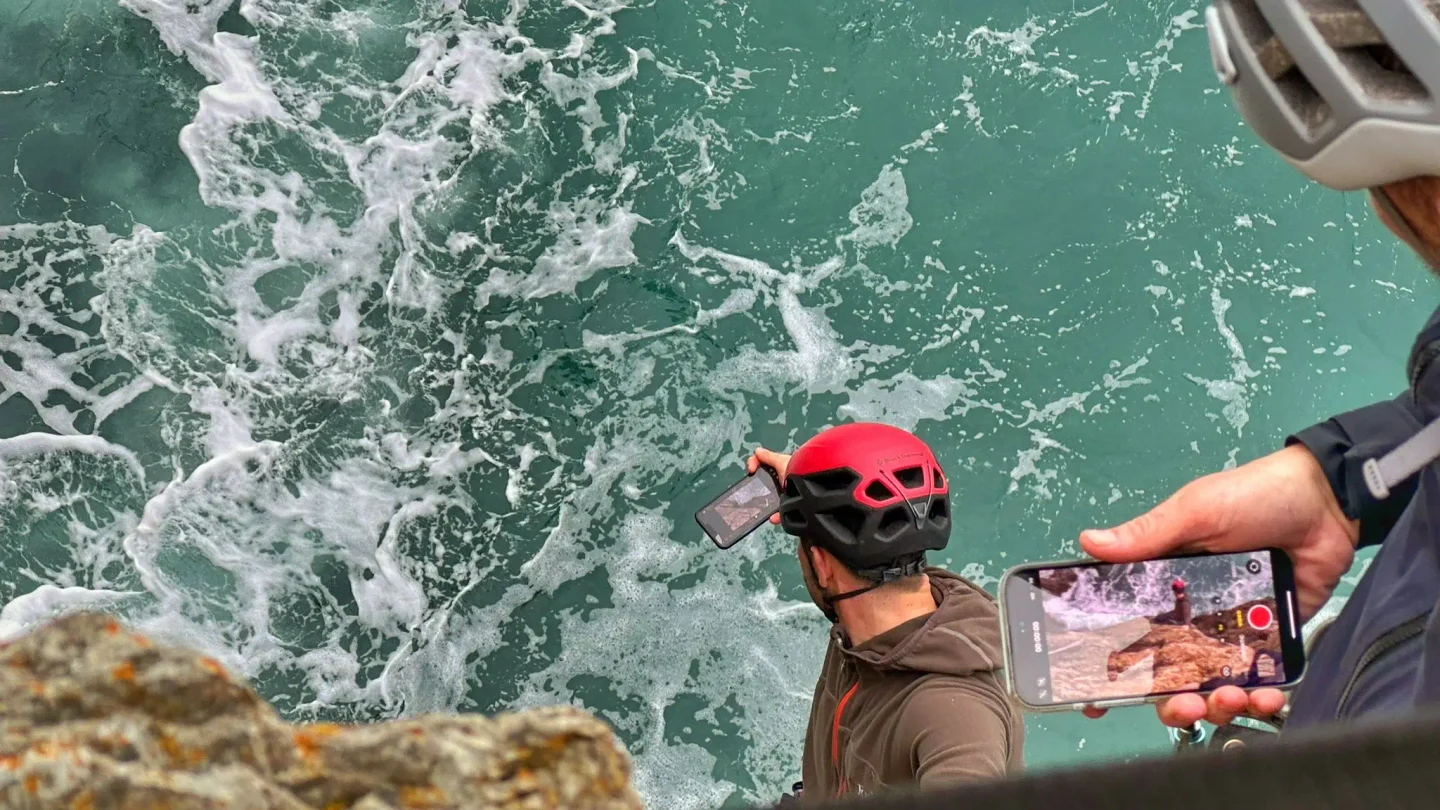 Crew member in a climbing helmet on a clifftop, filming on a phone above rough sea as a colleague records him.