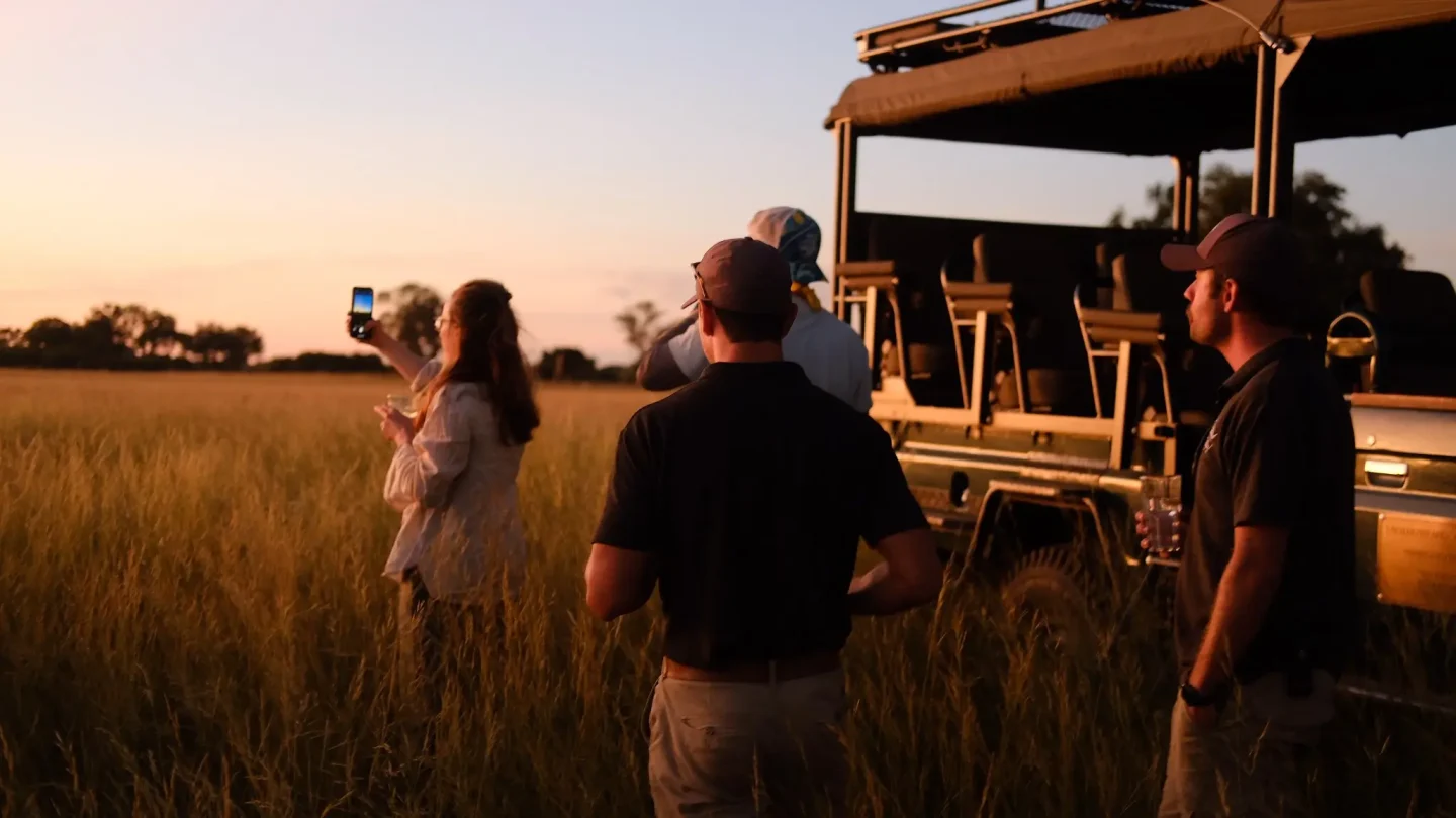 Film crew standing in long grass beside an open safari vehicle at sunset, one person taking a photo on a phone.