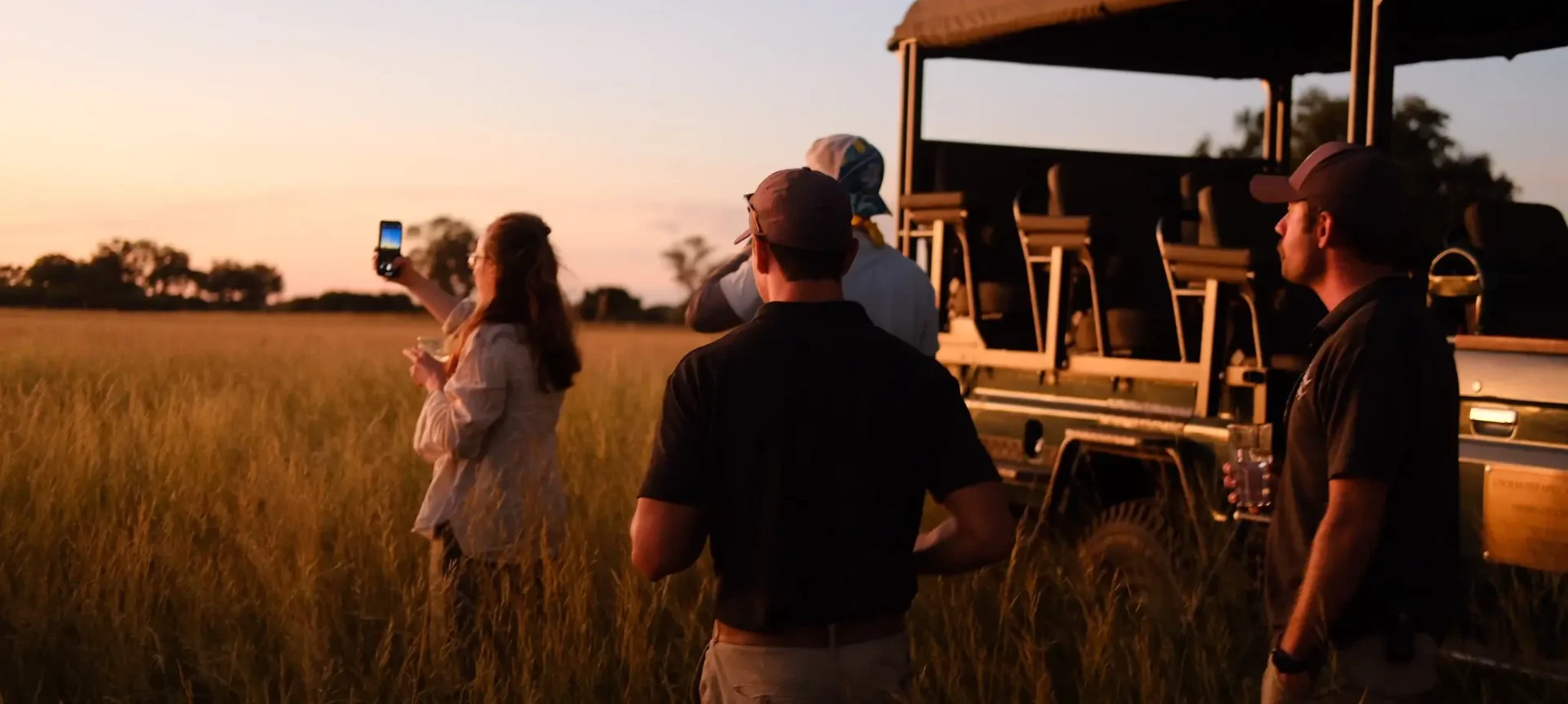 Film crew standing in long grass beside an open safari vehicle at sunset, one person taking a photo on a phone.
