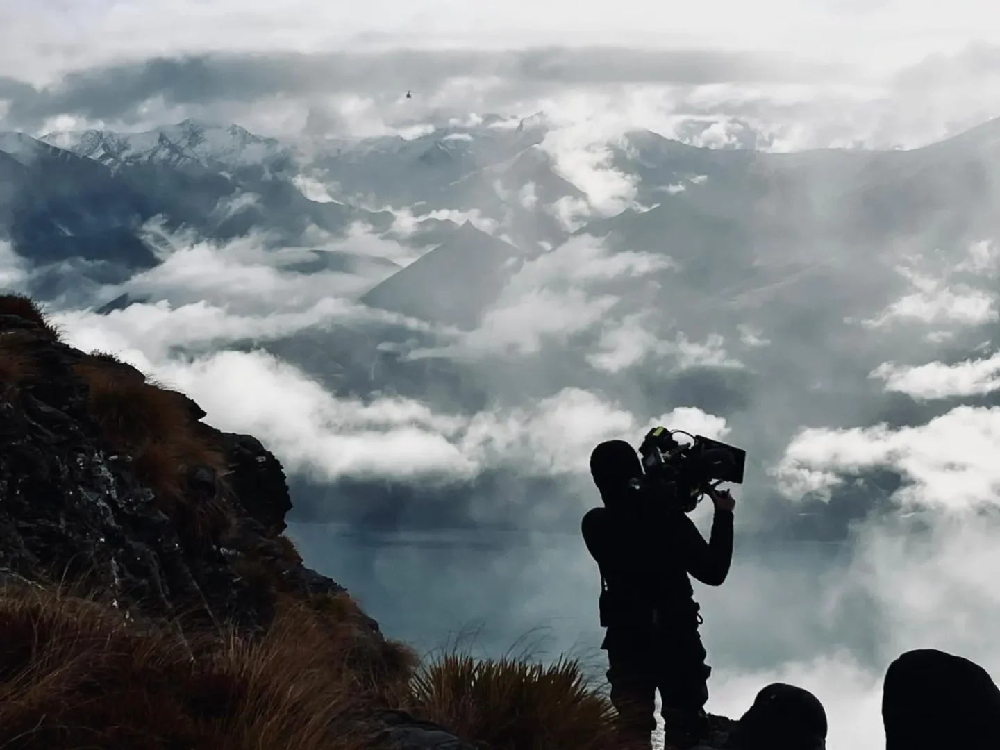 Silhouetted camera operator filming from a mountain ridge above low cloud and snow-capped peaks.